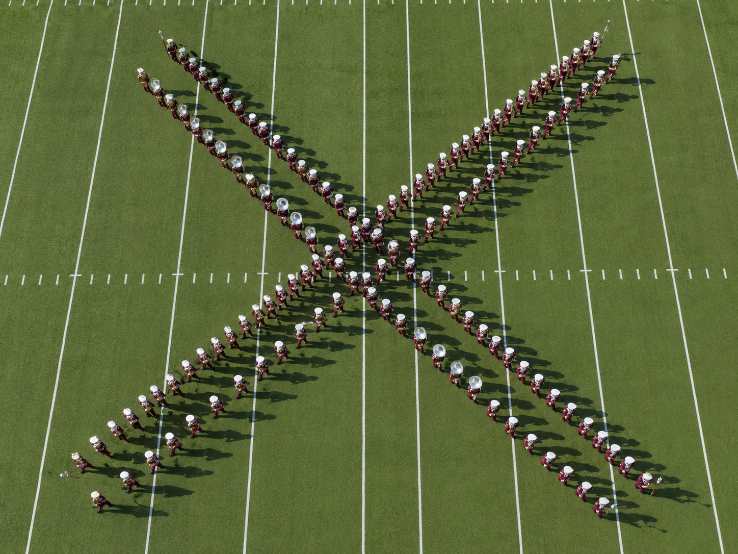 Aerial overhead view of a military-style marching band in maroon and white cadet uniforms with white pith helmets executing the criss-cross formation, two columns interlacing at a perfect 90-degree angle on a football field