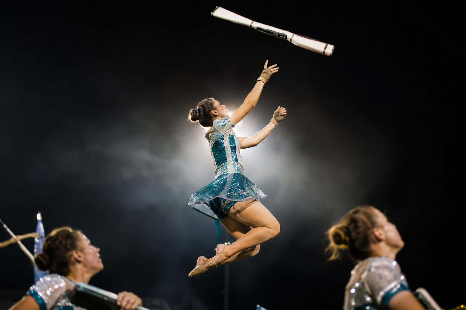 High school color guard performer mid-air during a rifle toss, sequined teal-and-silver costume catching stadium light, rifle frozen at the apex of its spin against a darkened night sky, fellow guard members slightly blurred in the foreground