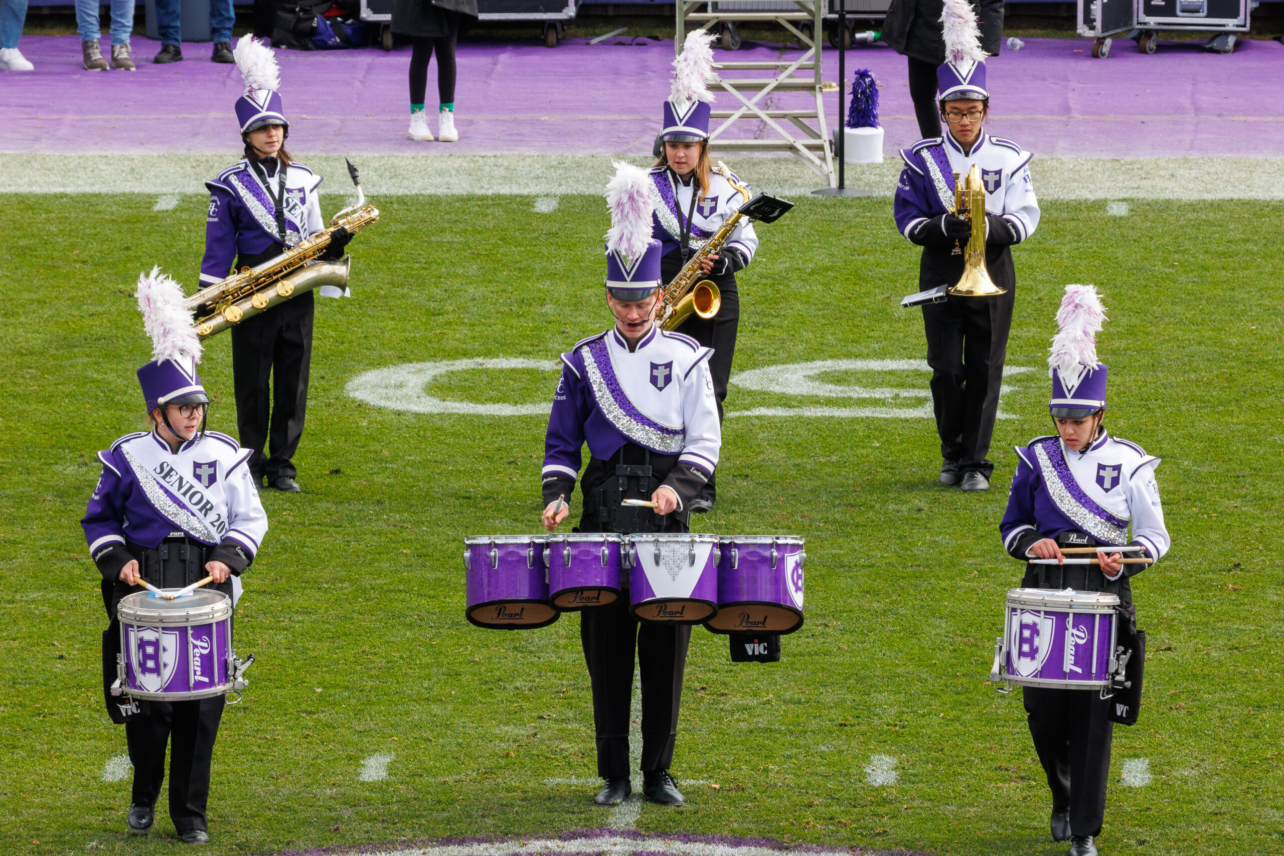 The College of the Holy Cross Goodtime Marching Band performing on Fitton Field in purple and white uniforms, brass instruments visible in mid-performance