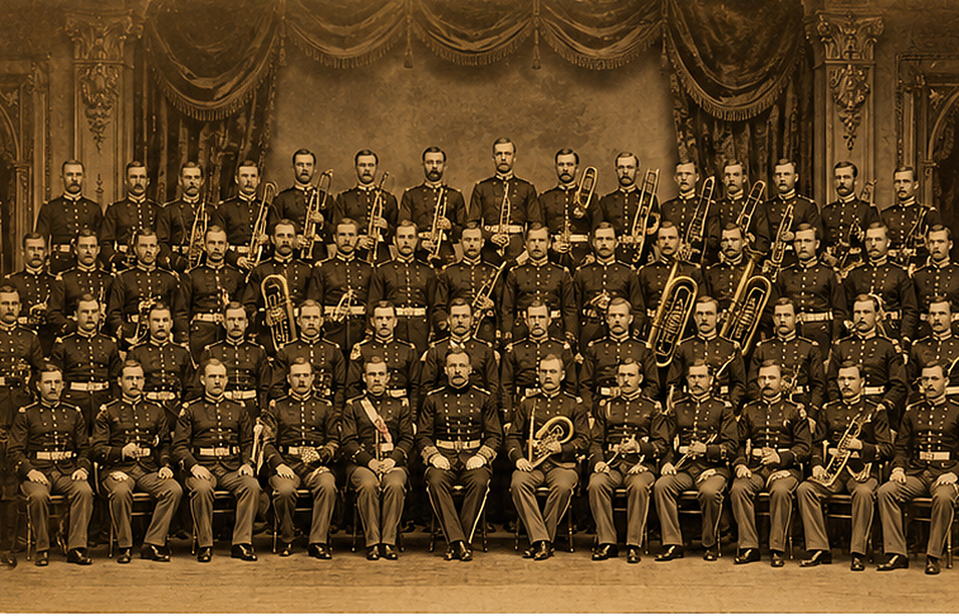 Sepia-toned vintage group photograph of approximately fifty members of the United States Marine Band c. 1890, in formal Marine dress-blue uniforms with brass instruments held vertically in front, arranged in three tiered rows against an ornate concert-hall backdrop with heavy drapes