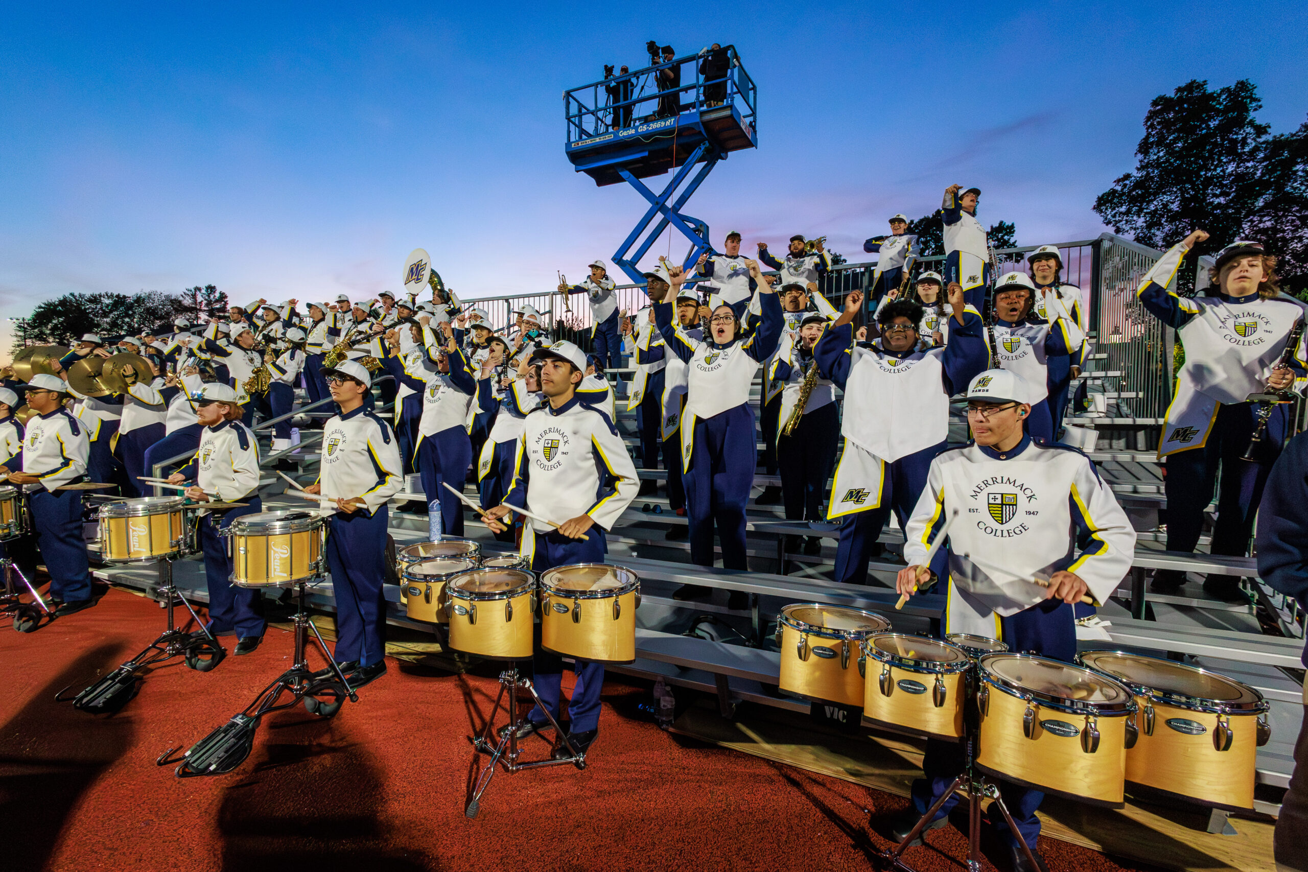 Merrimack College marching band in blue and gold uniforms playing brass instruments at a night football game under stadium lights