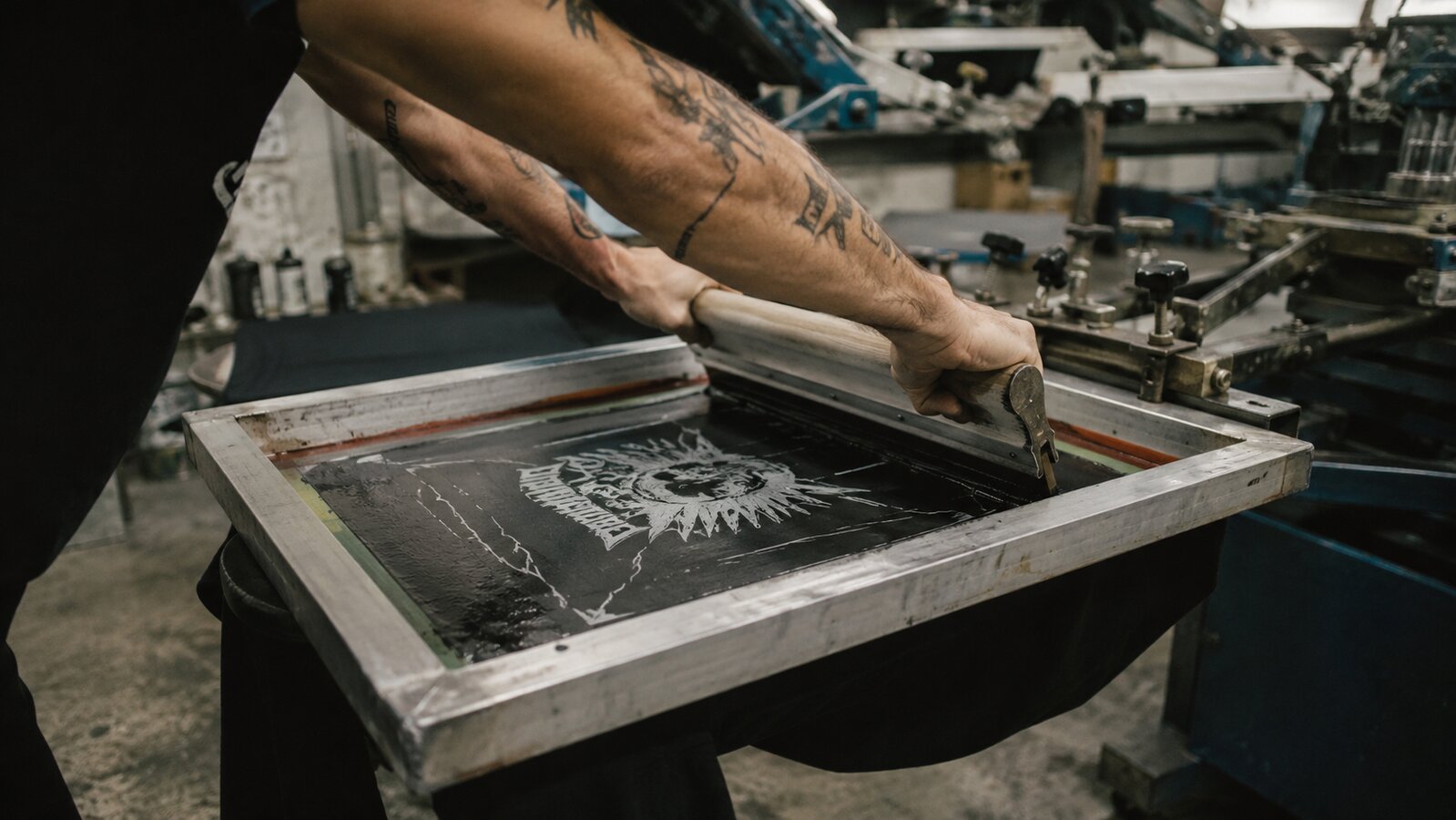 Screen-printer pulling ink across a band-name design on a t-shirt platen