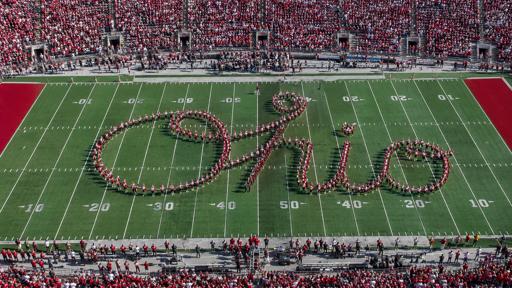 Aerial overhead view of The Ohio State University Marching Band forming 'Ohio' in flowing cursive script across the 50-yard line in front of a packed stadium of scarlet-and-gray fans