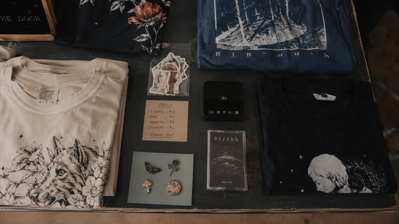 Tour merch table flat-lay with tees, enamel pins, cassette, and stickers